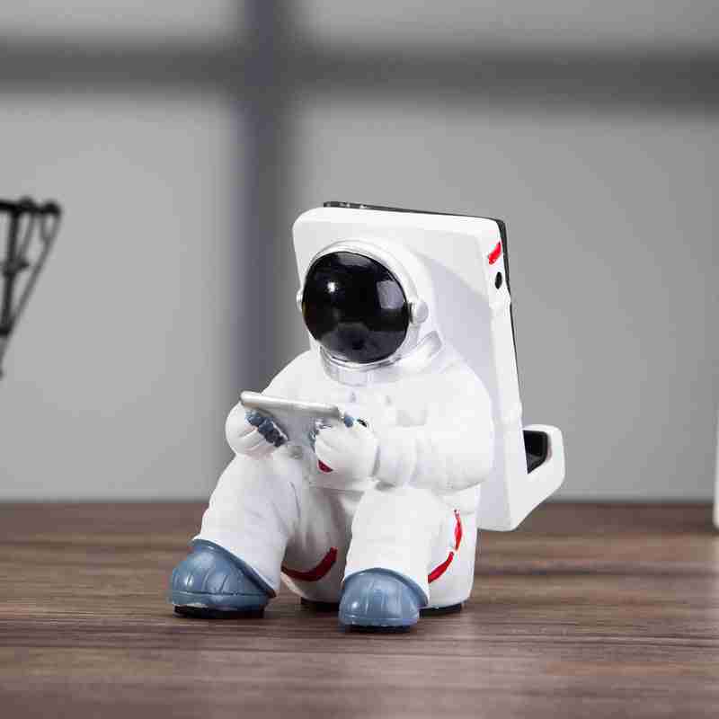 Astronaut Mobile Phone Stand Fun & Functional Desk Holder made of resin, featuring a seated astronaut figure holding a remote and supporting a smartphone.
